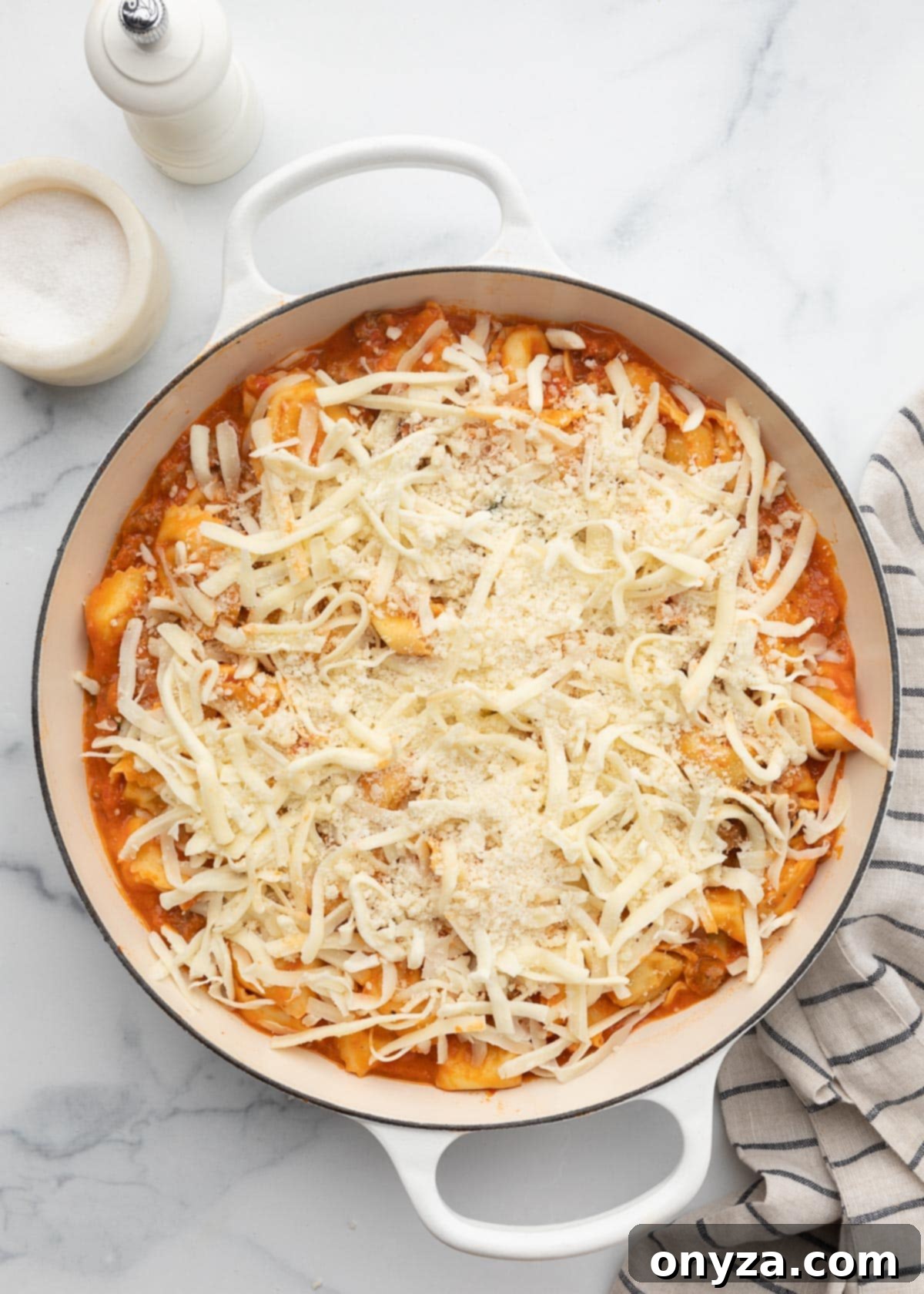 Unbaked cheese-topped tortellini casserole in a white enameled cast iron pan, glistening and ready for the oven.