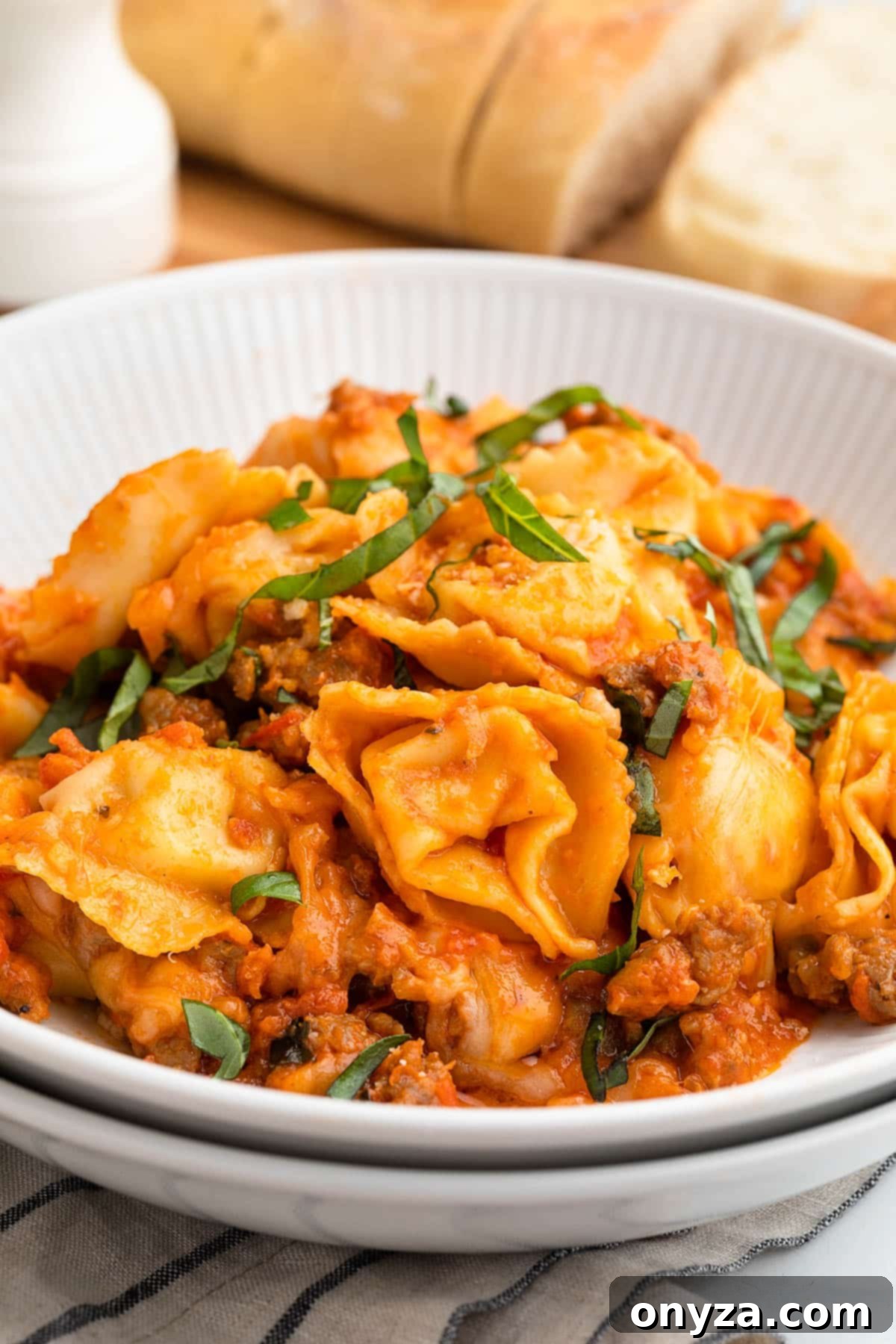 Baked tortellini with sausage served in a double-stacked white pasta bowl, garnished with delicate julienned basil leaves.