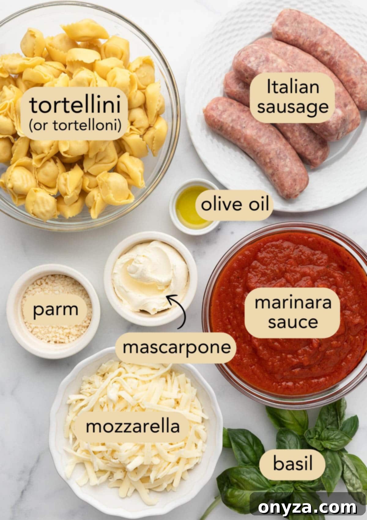 Overhead view of all ingredients needed to make baked tortellini with sausage, neatly arranged in bowls on a white marble board with text labels.
