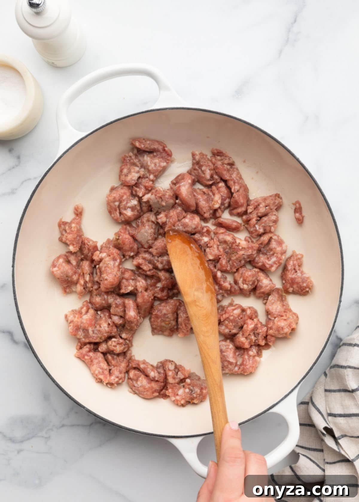Raw Italian sausage in a white enameled cast iron pan being stirred with a wooden spoon, breaking it into crumbles.