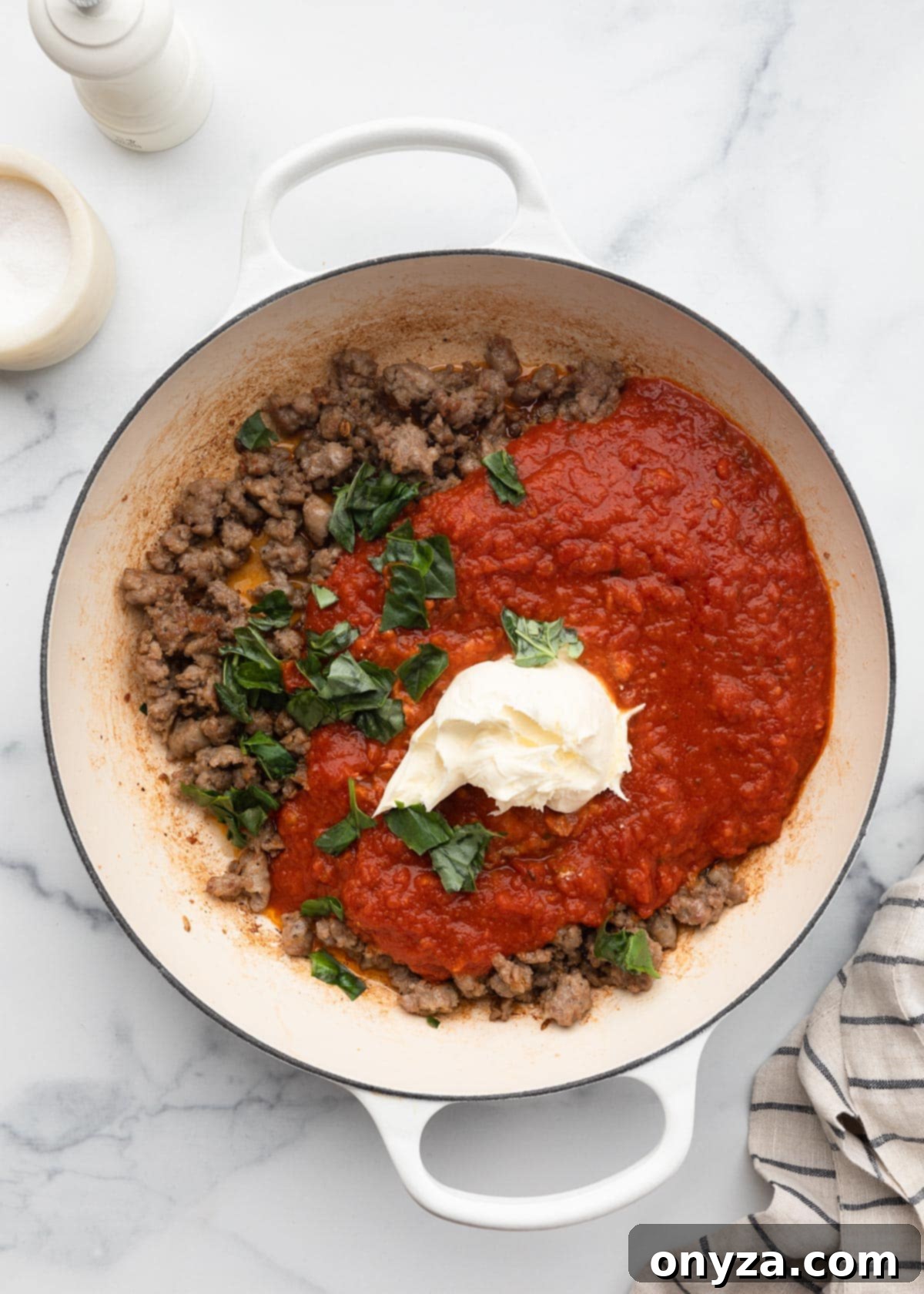 Marinara sauce, browned crumbled sausage, mascarpone cheese, and torn basil leaves combining in a white enameled cast iron pot.