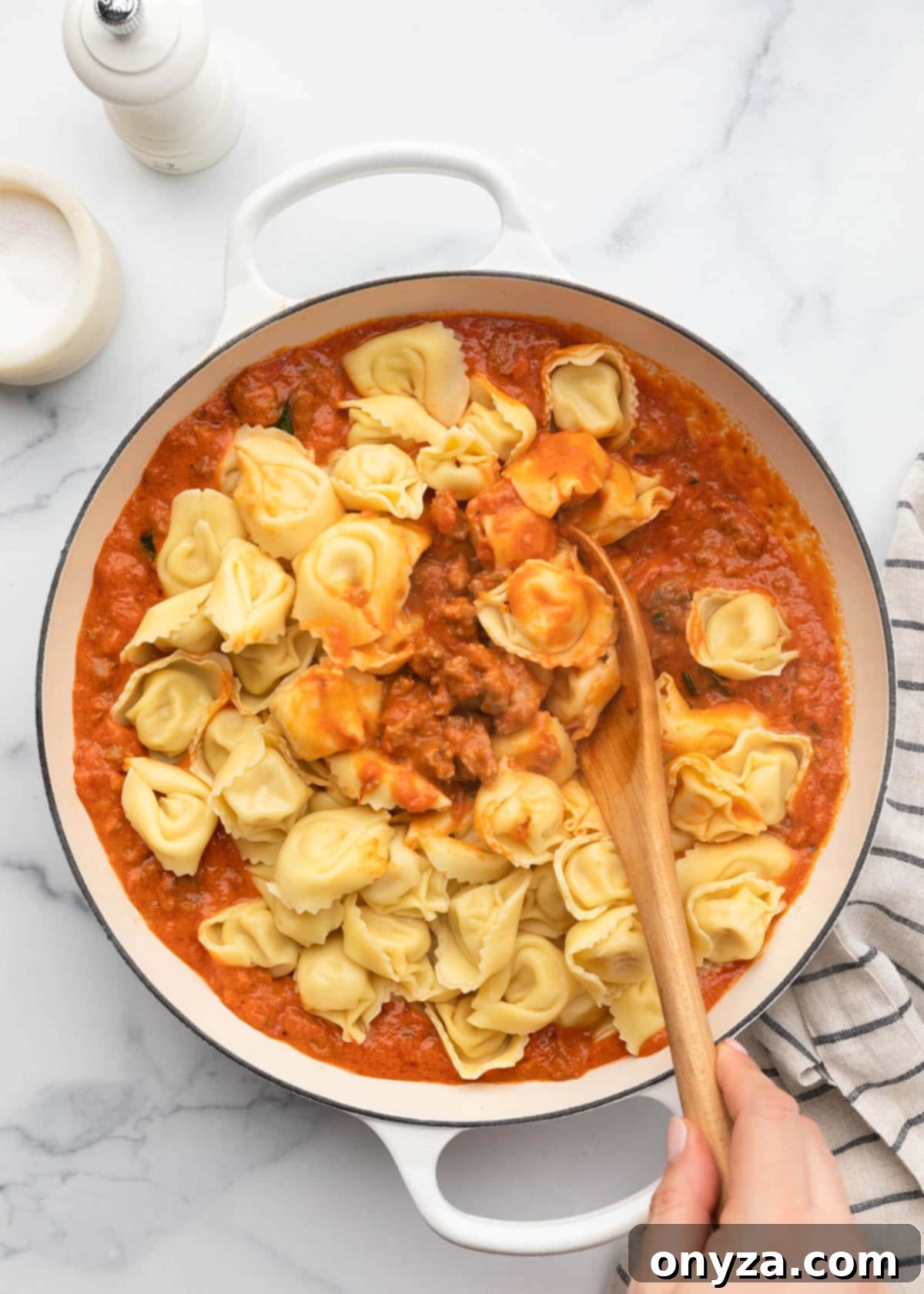 Stirring cooked tortellini into the savory sausage marinara sauce with a wooden spoon in a white enameled cast iron pan.
