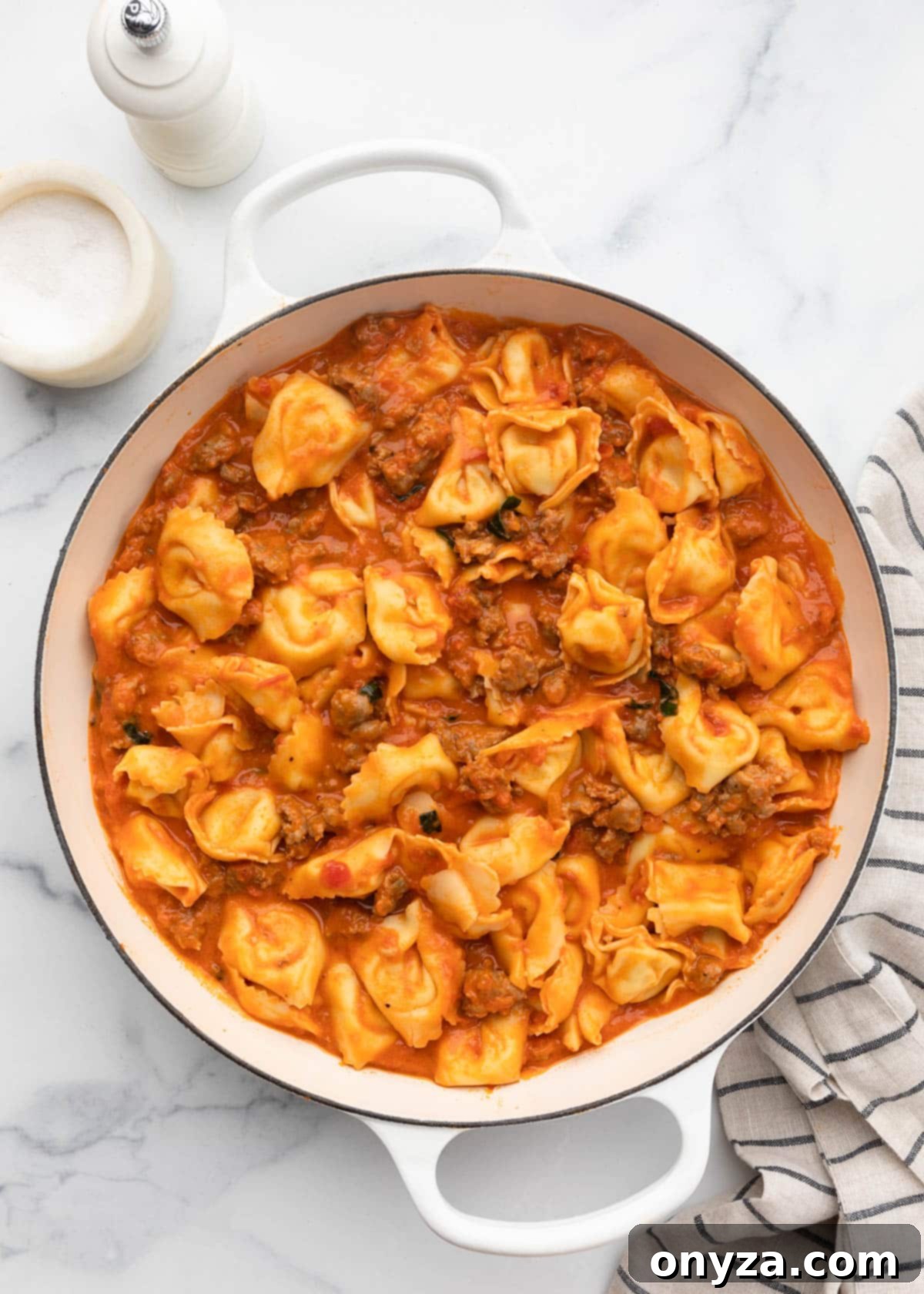 Cooked tortellini stirred into a rich sausage tomato sauce in a white enameled cast iron pan, ready for the cheese topping.