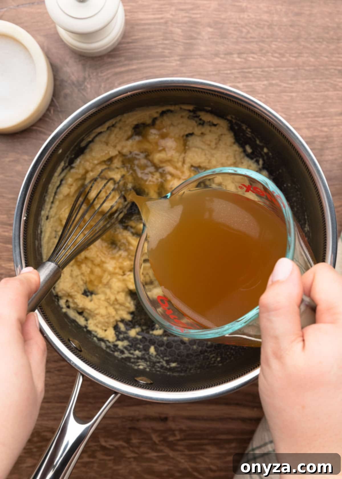 chicken stock being poured from a measuring cup into a pot of roux while being whisked