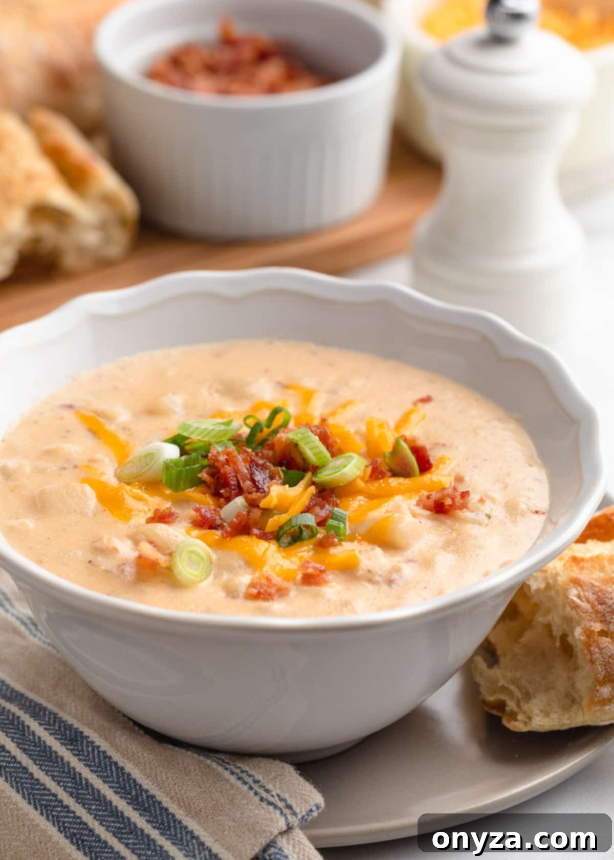 A steaming bowl of Crockpot Hashbrown Potato Soup, garnished with fresh chives and bacon, resting on a rustic wooden board next to a freshly baked baguette and a white pepper mill.