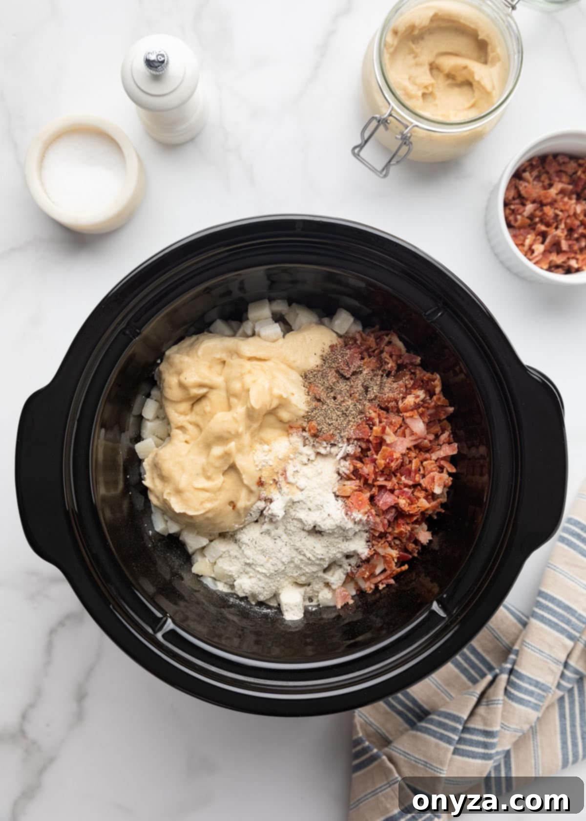 Overhead shot of frozen hash browns, condensed cream of chicken soup, crumbled bacon, and various seasonings nestled in a slow cooker, ready for stirring on a white marble surface.