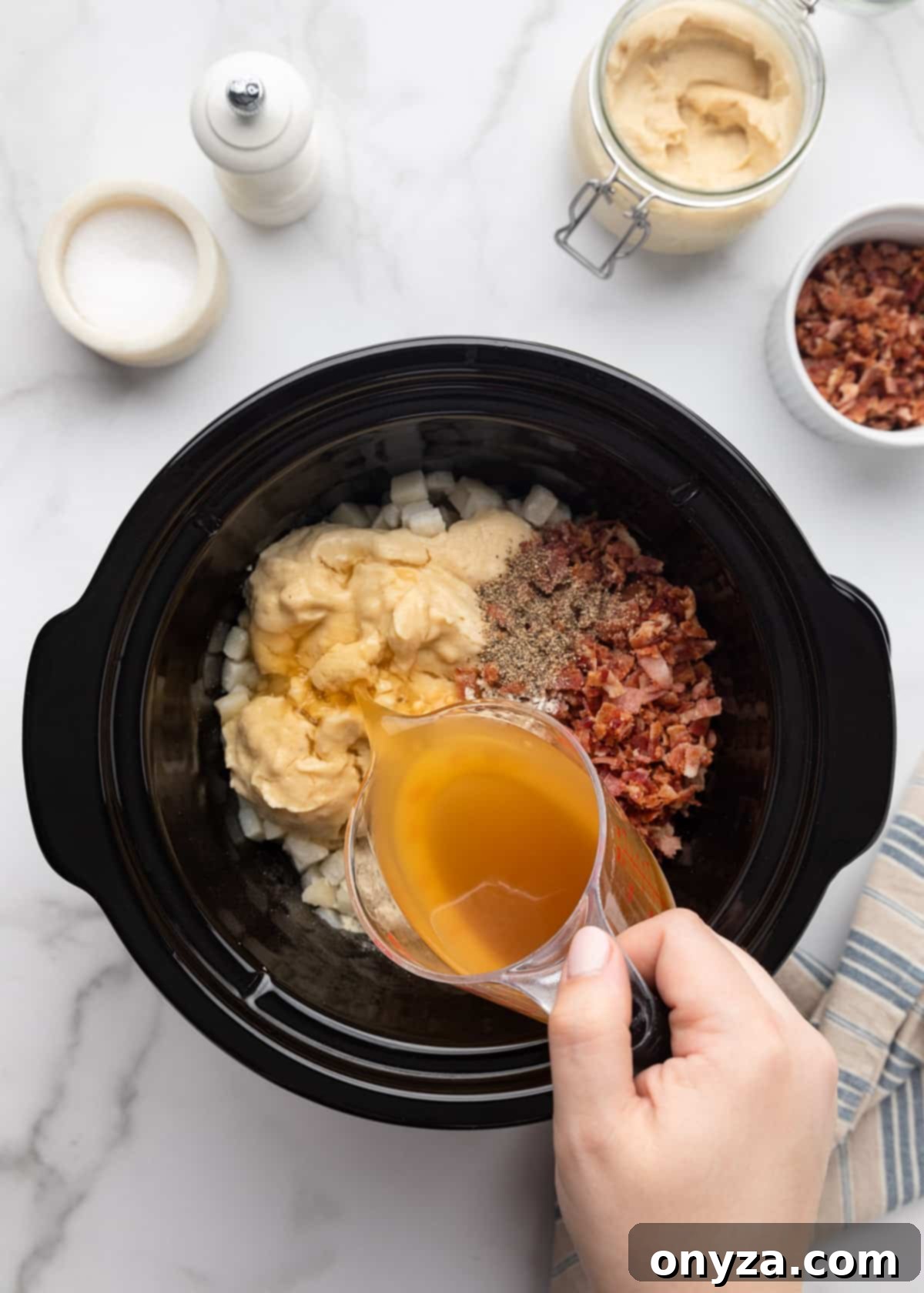 Chicken stock being carefully poured from a clear measuring cup into the slow cooker, which is already filled with hash brown potato soup ingredients on a white marble board.