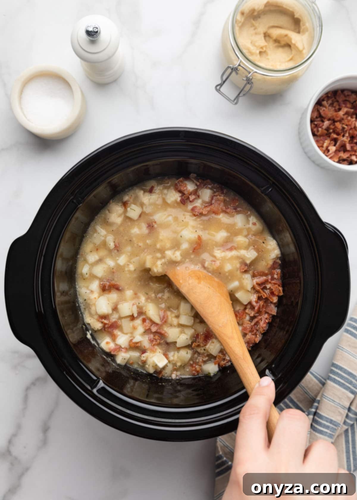 Overhead shot of a wooden spoon gently stirring the hash brown potato soup ingredients within the slow cooker, ensuring everything is well combined on a white marble background.