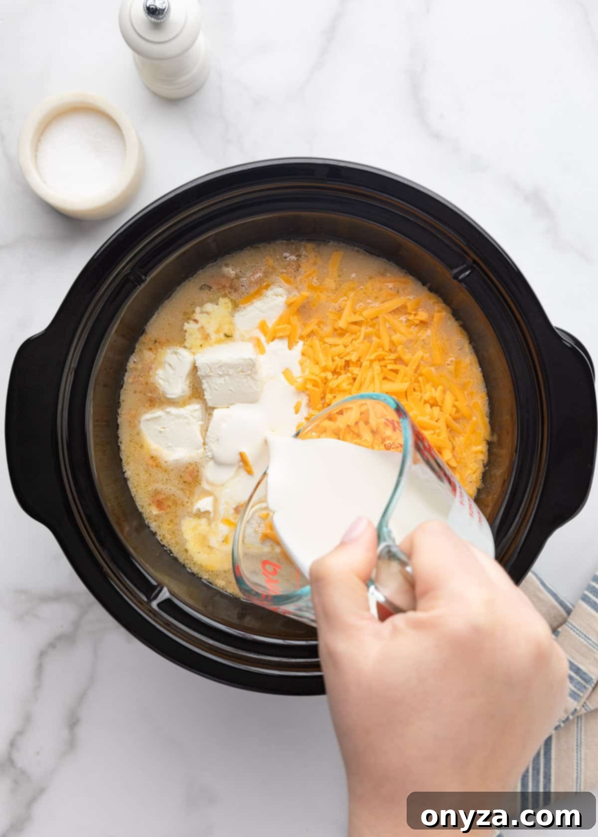 Heavy cream being poured from a measuring cup into the slow cooker, which already contains the partially cooked potato soup, cream cheese cubes, and shredded cheddar cheese, all on a white marble board.
