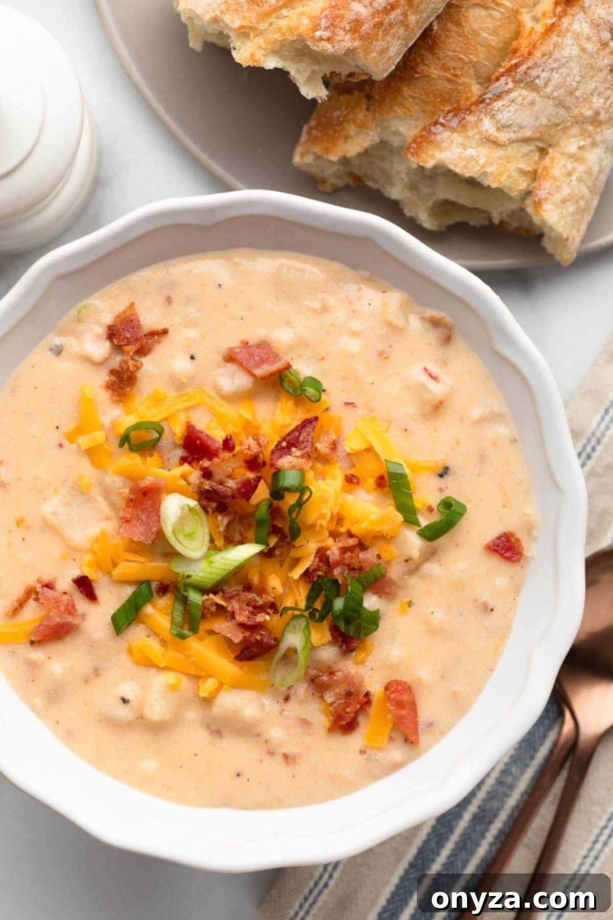 An inviting overhead shot of a white bowl filled with creamy Crockpot Hashbrown Potato Soup, accompanied by a white pepper mill and golden wedges of baguette, ready to be enjoyed.
