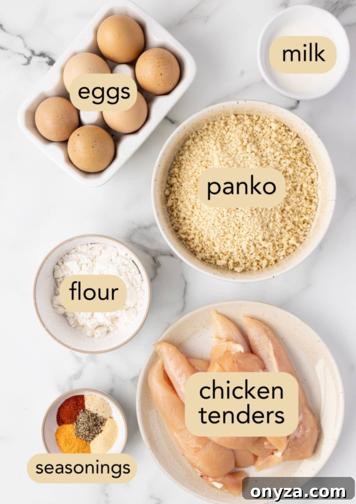 Overhead view of air fryer chicken tender ingredients neatly arranged in bowls on a white marble surface, including flour, egg wash, and panko breadcrumbs.