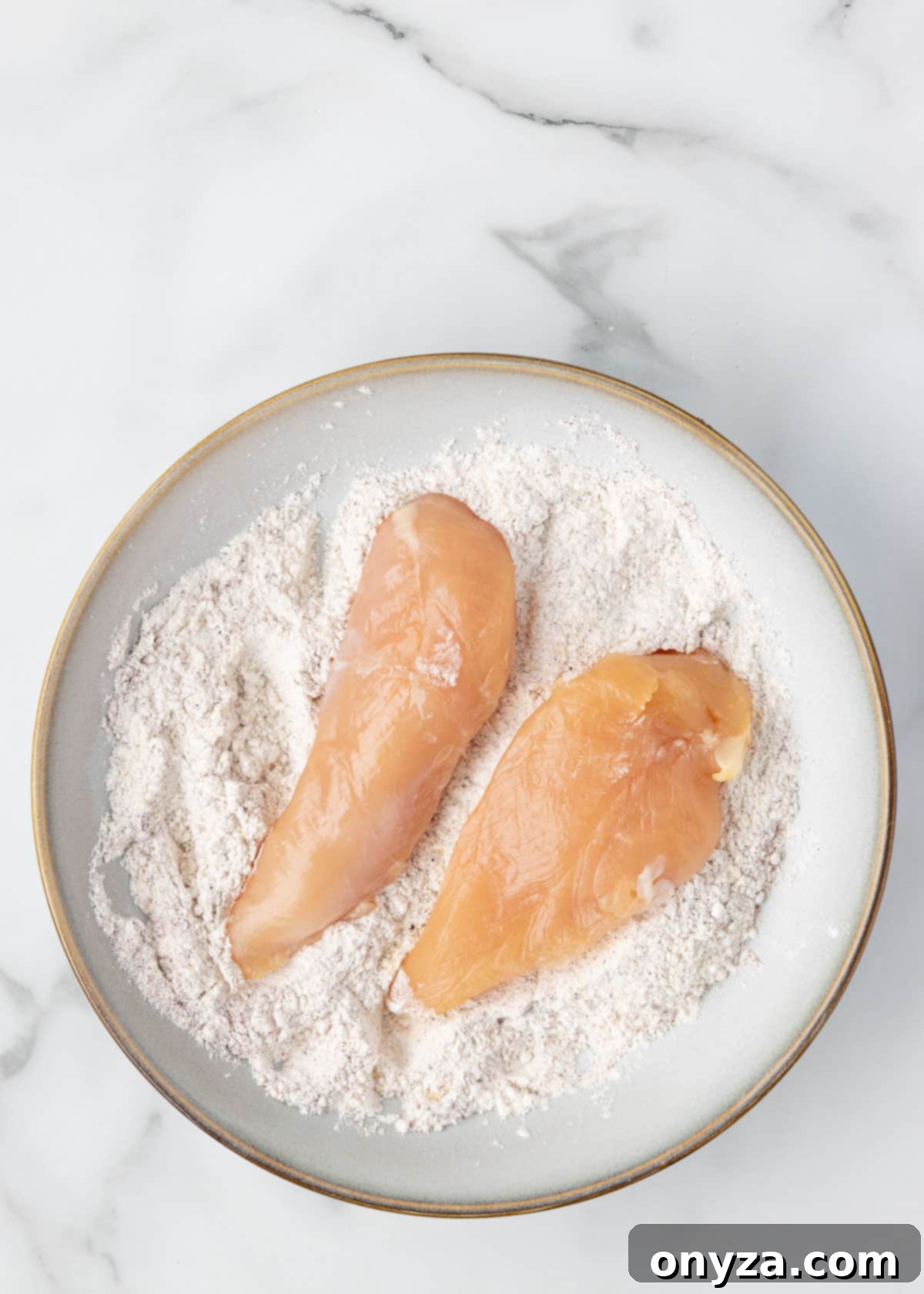 Raw chicken tenders lightly coated in seasoned flour in a shallow plate on a white marble board, ready for the next step.