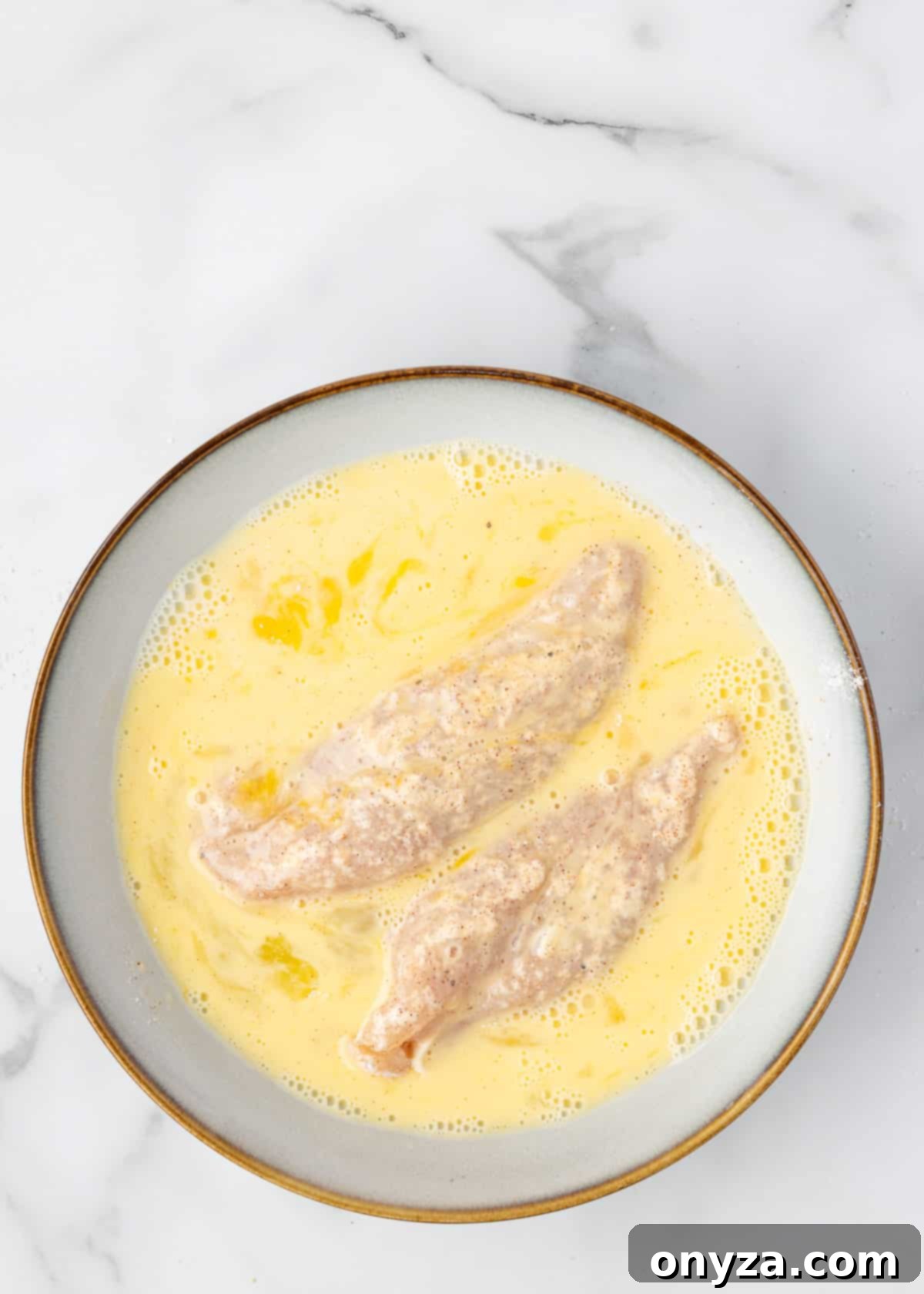 Flour-dredged chicken tenders being dipped into a bowl of whisked egg wash, with excess dripping off, on a white marble board.