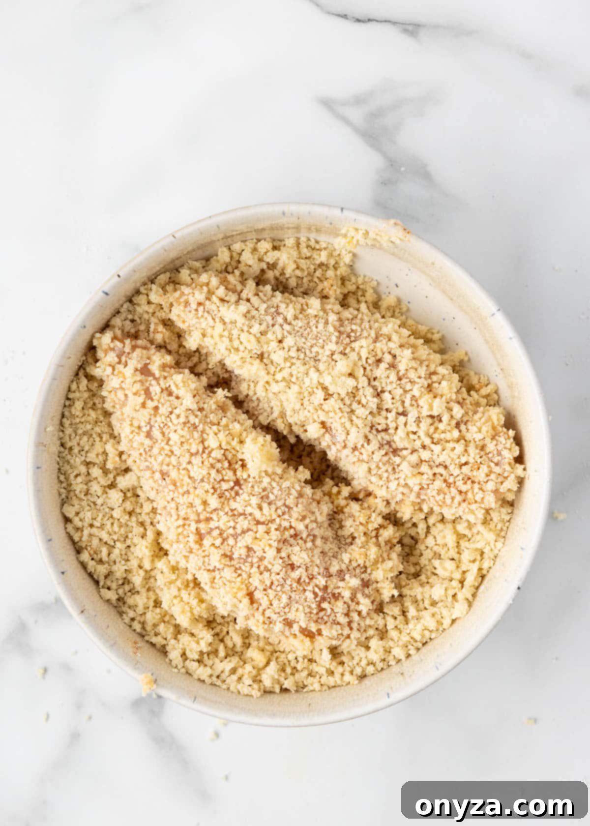 Raw chicken tenders fully coated with a generous layer of seasoned panko bread crumbs, neatly arranged on a white marble board, ready for air frying.