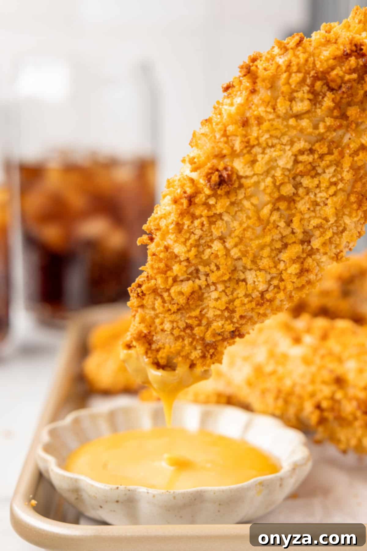 A golden brown air fryer chicken tender being dipped into a small bowl of creamy honey mustard sauce, with a glass of refreshing cola in the soft background.