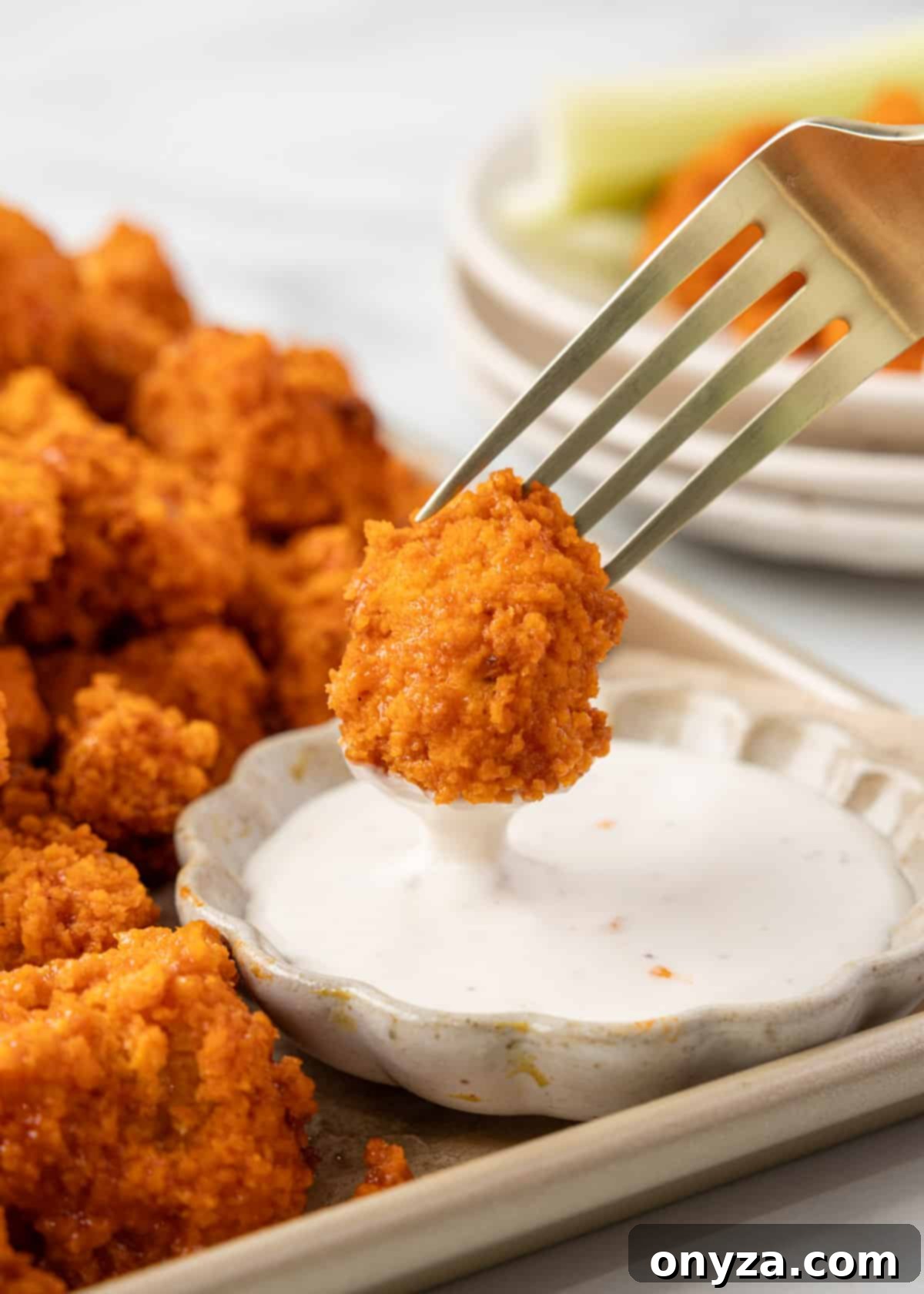 air fryer buffalo chicken bite being dipped into a bowl of ranch dressing from a fork. A pile of chicken bites surrounds the bowl on a rimmed baking sheet.