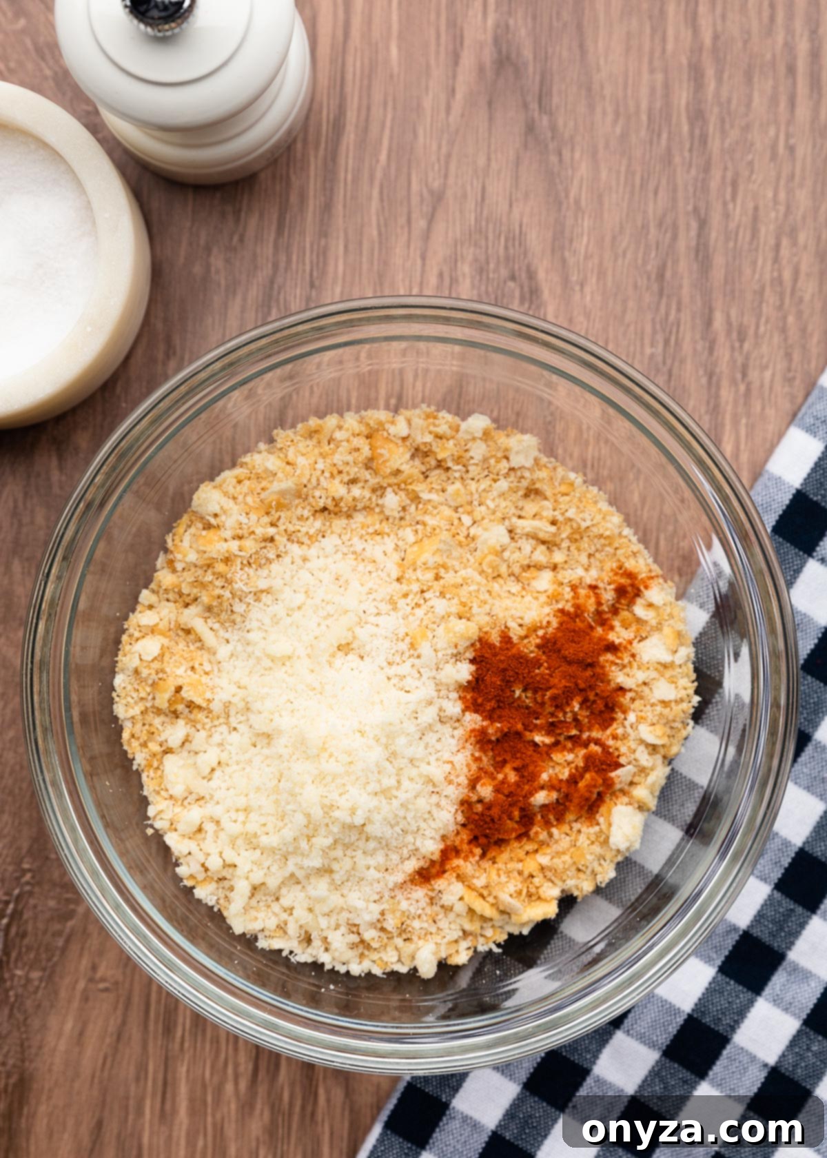 Crushed Ritz crackers in a bowl alongside Parmesan cheese and paprika on a wooden board next to a blue and white checkered napkin, ingredients for the casserole topping.
