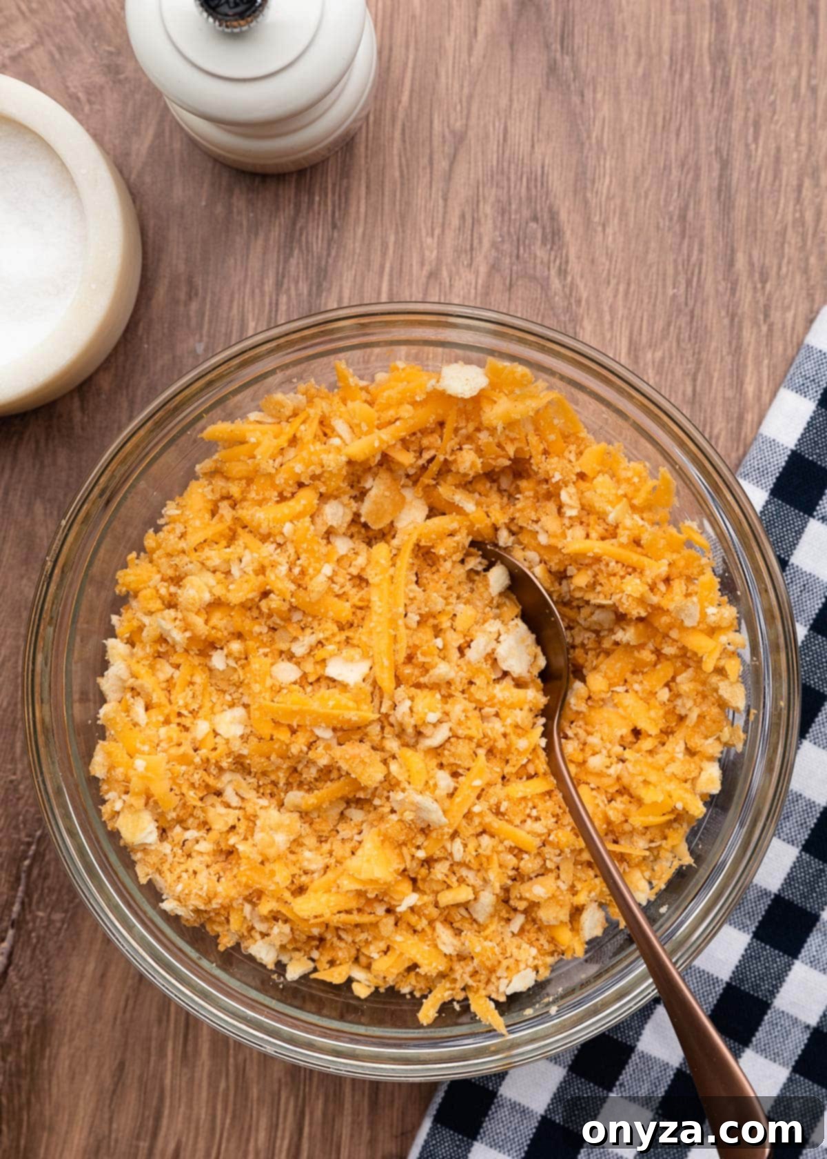 A bowl of crushed cracker topping, mixed with melted butter and shredded cheddar, resting on a wooden board next to a blue and white checkered napkin.