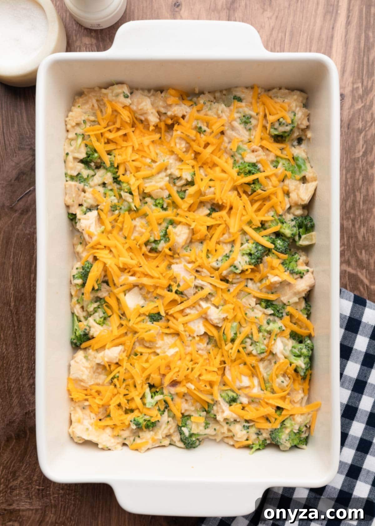 Half of the chicken broccoli rice casserole layered into a white baking dish, generously sprinkled with shredded cheddar cheese, ready for the next layer.