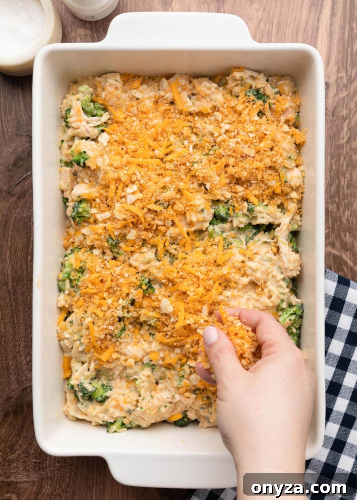Sprinkling shredded cheddar and the crushed cracker crumb topping over the chicken broccoli rice casserole in a white baking dish, preparing it for baking.