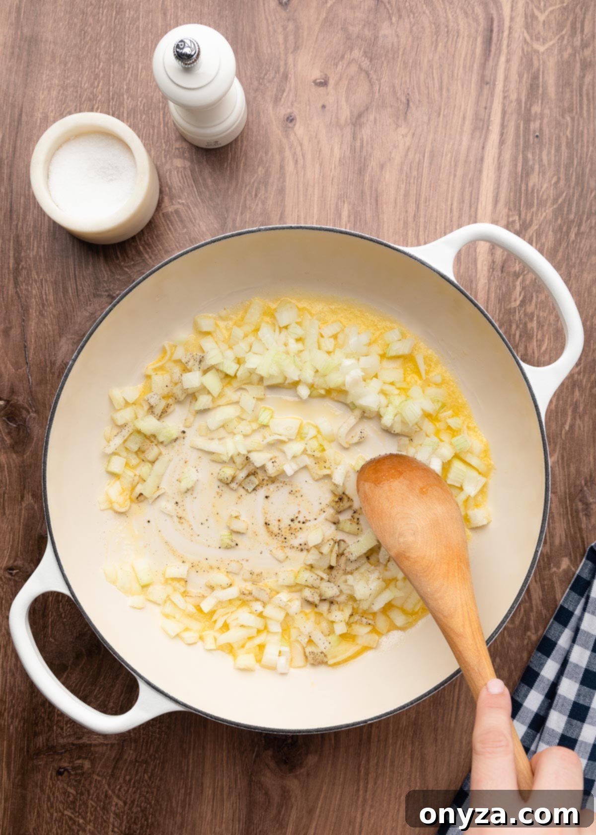 Sautéing chopped yellow onions in melted butter with salt and pepper in a white enameled cast iron pot, stirred with a wooden spoon.