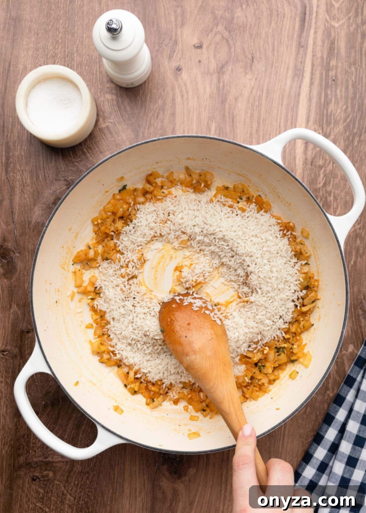 Stirring uncooked extra-long grain white rice into sautéed seasoned onions in a white enameled cast iron pot with a wooden spoon.