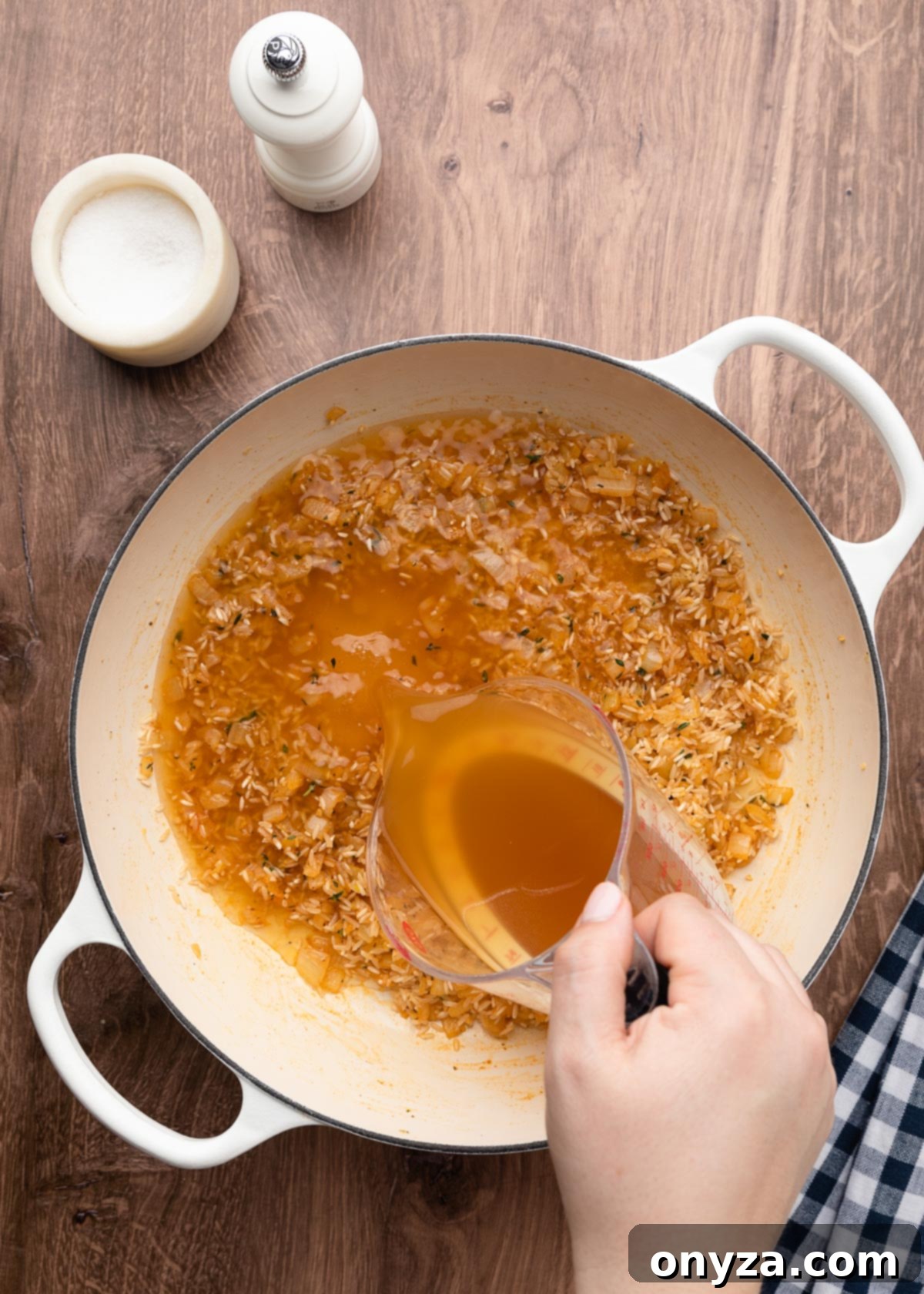 Pouring chicken stock from a liquid measuring cup into a white enameled cast iron pot containing seasoned uncooked rice, forming the base of the casserole.