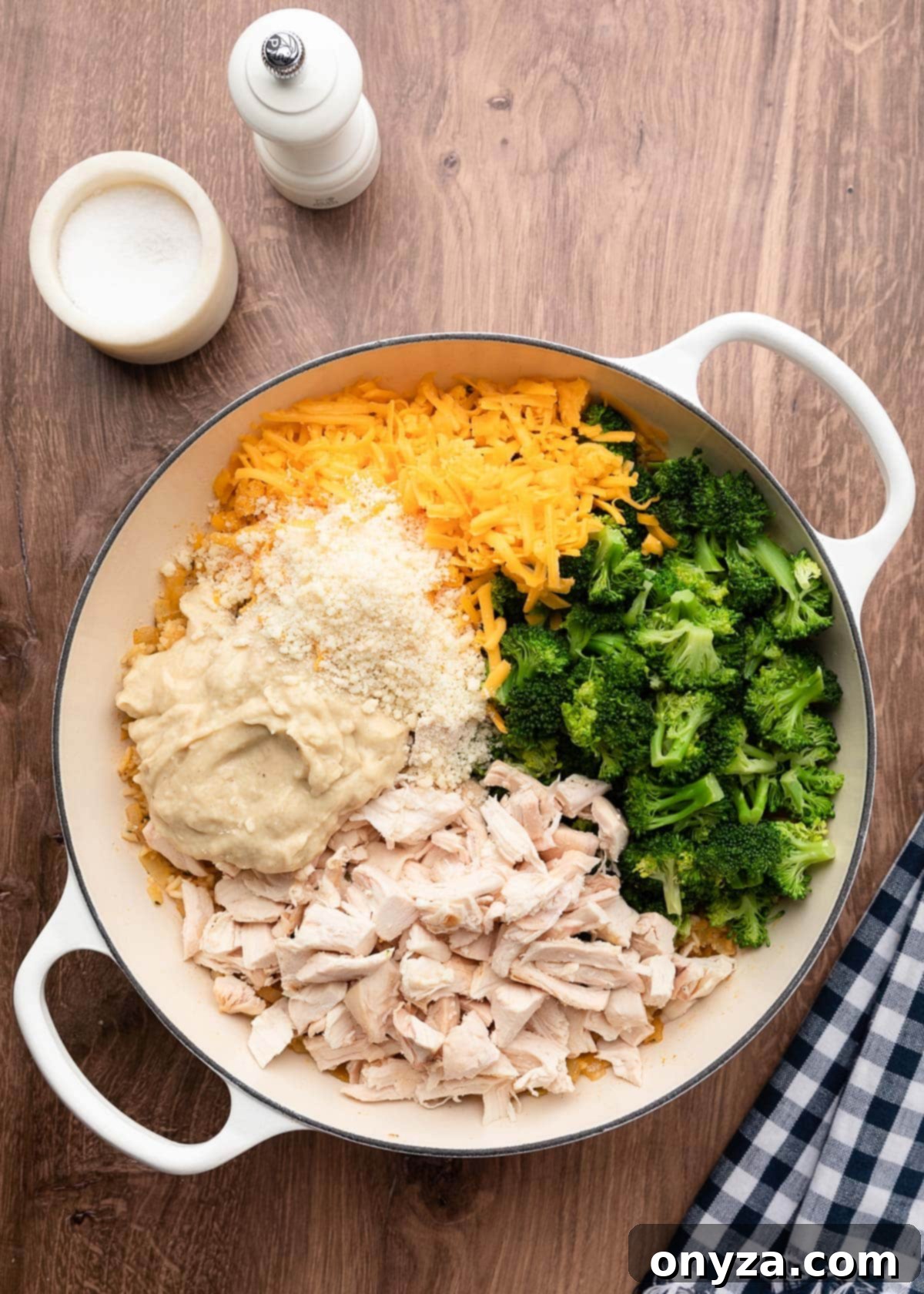 Ingredients for the casserole base, including condensed cream of chicken soup, shredded cheddar, cooked broccoli florets, Parmesan cheese, and cooked chicken breast, layered over the cooked rice in a white enameled cast iron pot.
