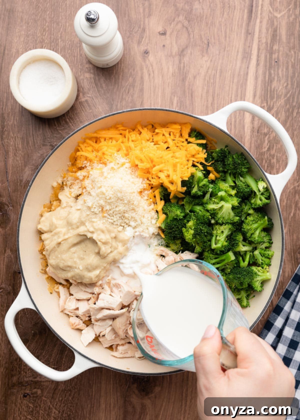Pouring half and half from a liquid measuring cup into a pot filled with the unmixed chicken, broccoli, and rice casserole ingredients, ready to be combined.