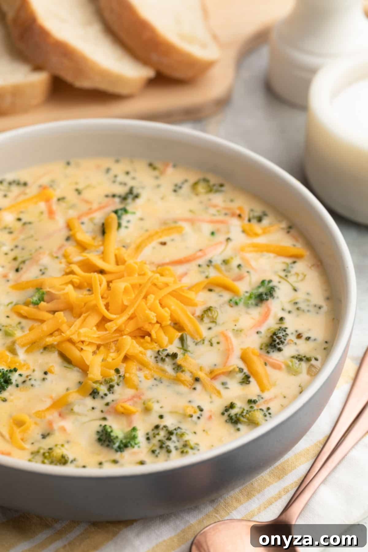 Broccoli Cheddar Soup in a blue and ivory ceramic bowl next to bronze soup spoons, a yellow and white striped napkin, and sliced crusty bread