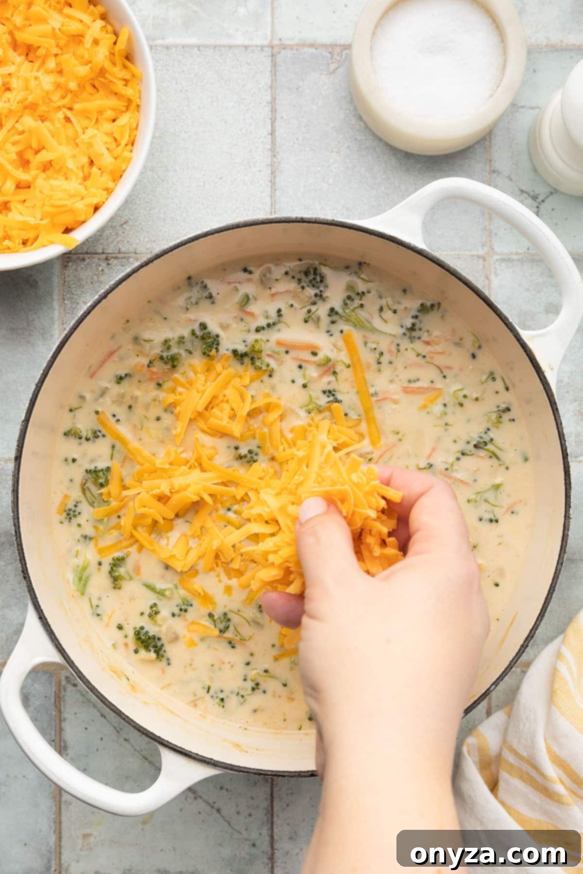adding shredded cheddar cheese to cooked broccoli cheddar soup in a white enameled cast iron dutch oven