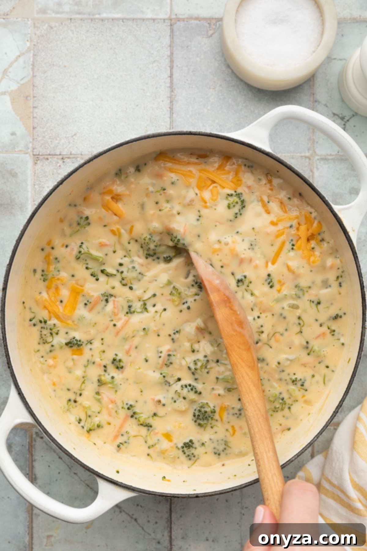 stirring cheddar cheese into a pot of broccoli cheese soup in a white enameled cast iron dutch oven with a wooden spoon