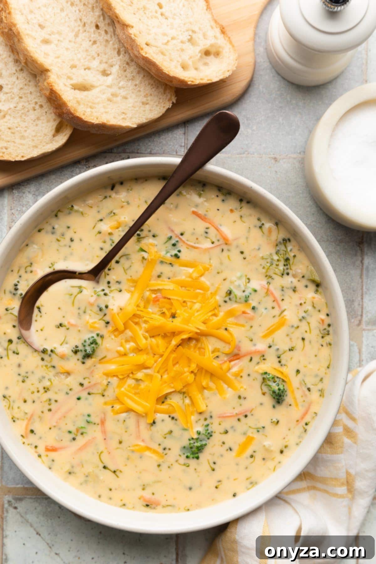 overhead of broccoli cheddar soup in an ivory ceramic bowl with a bronze soup spoon next to slices of crusty bread and a yellow and white striped napkin