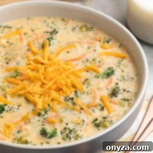 Broccoli Cheddar Soup in a blue and ivory ceramic bowl next to bronze soup spoons, a yellow and white striped napkin, and sliced crusty bread