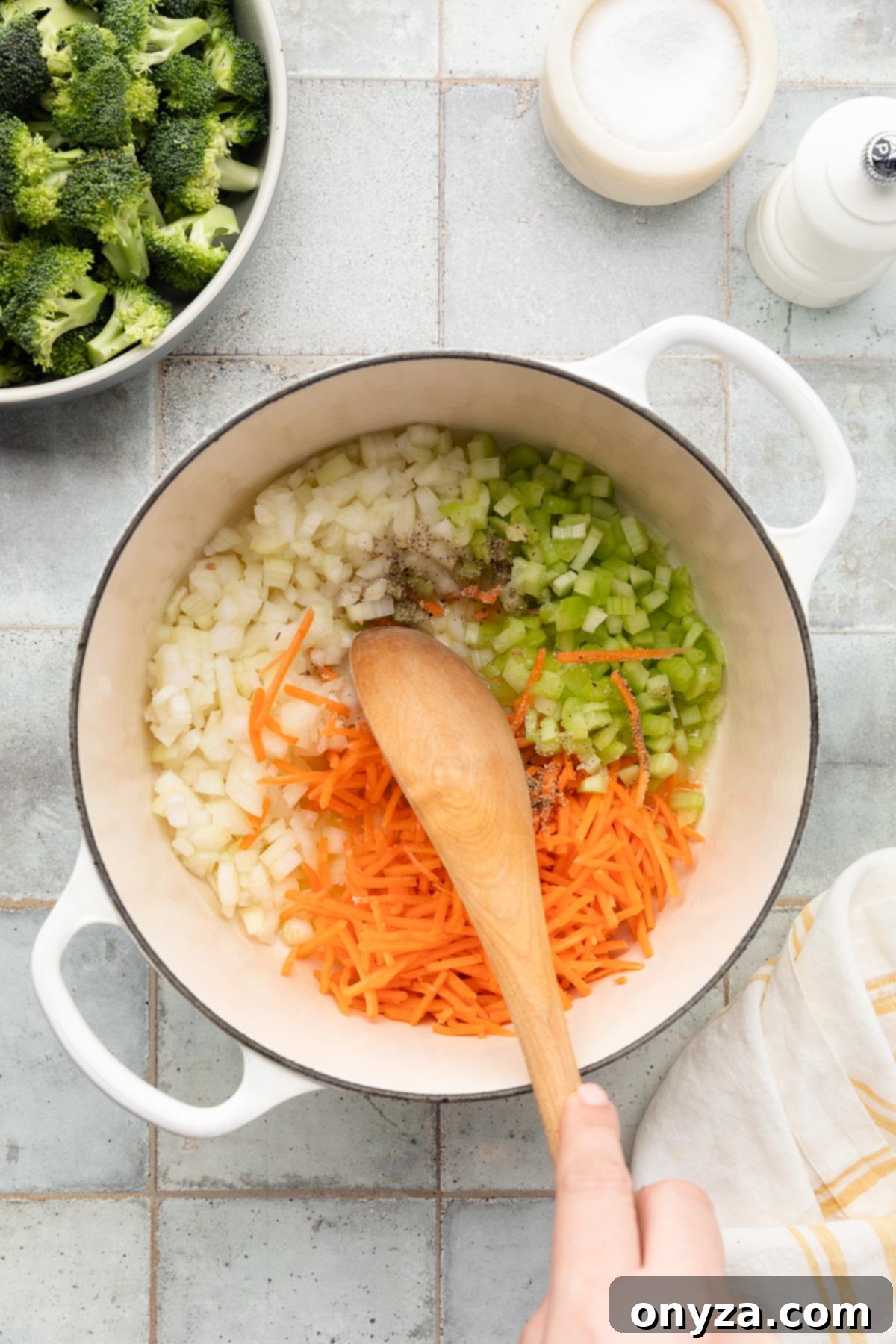 diced onion, diced celery, and shredded carrots being stirred in a white enameled cast iron dutch oven with a wooden spoon