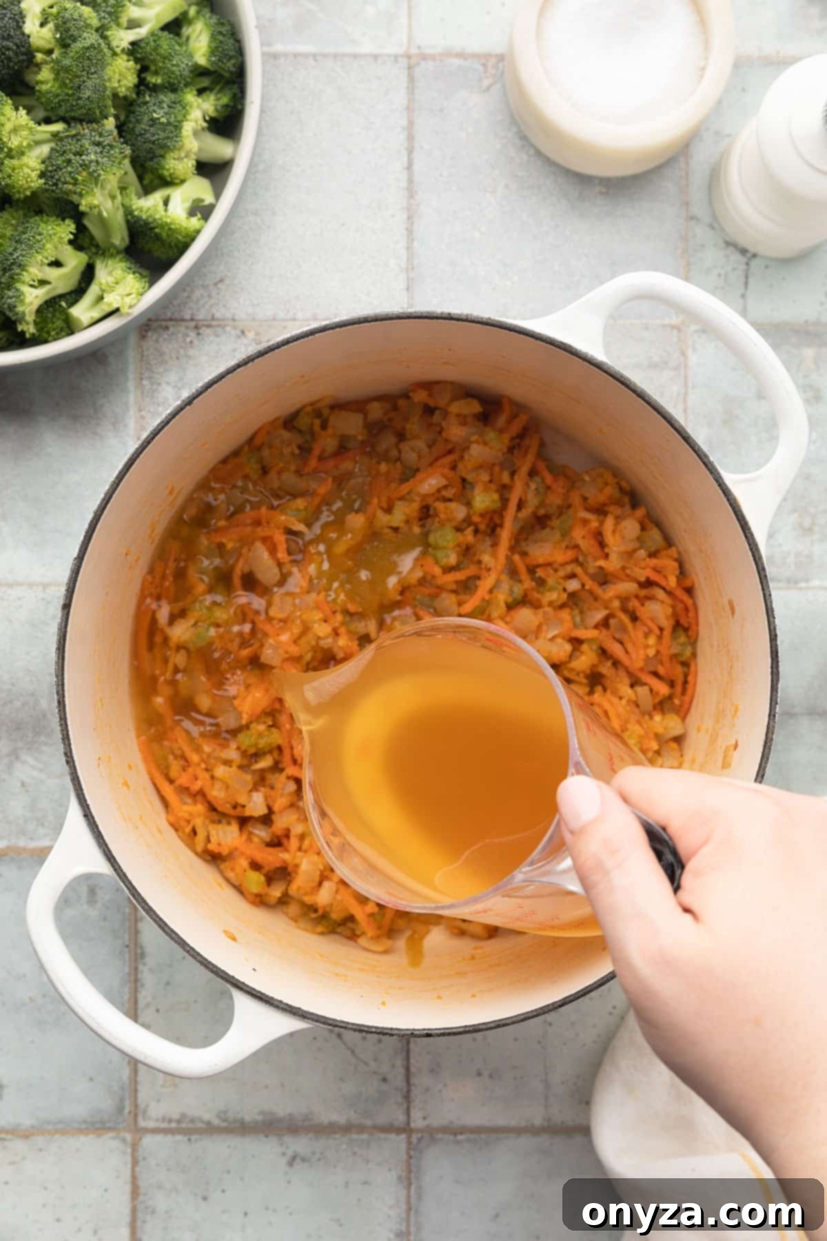 pouring chicken stock into a flour-coated sautéed mirepoix in a white enameled cast iron dutch oven from a liquid measuring cup