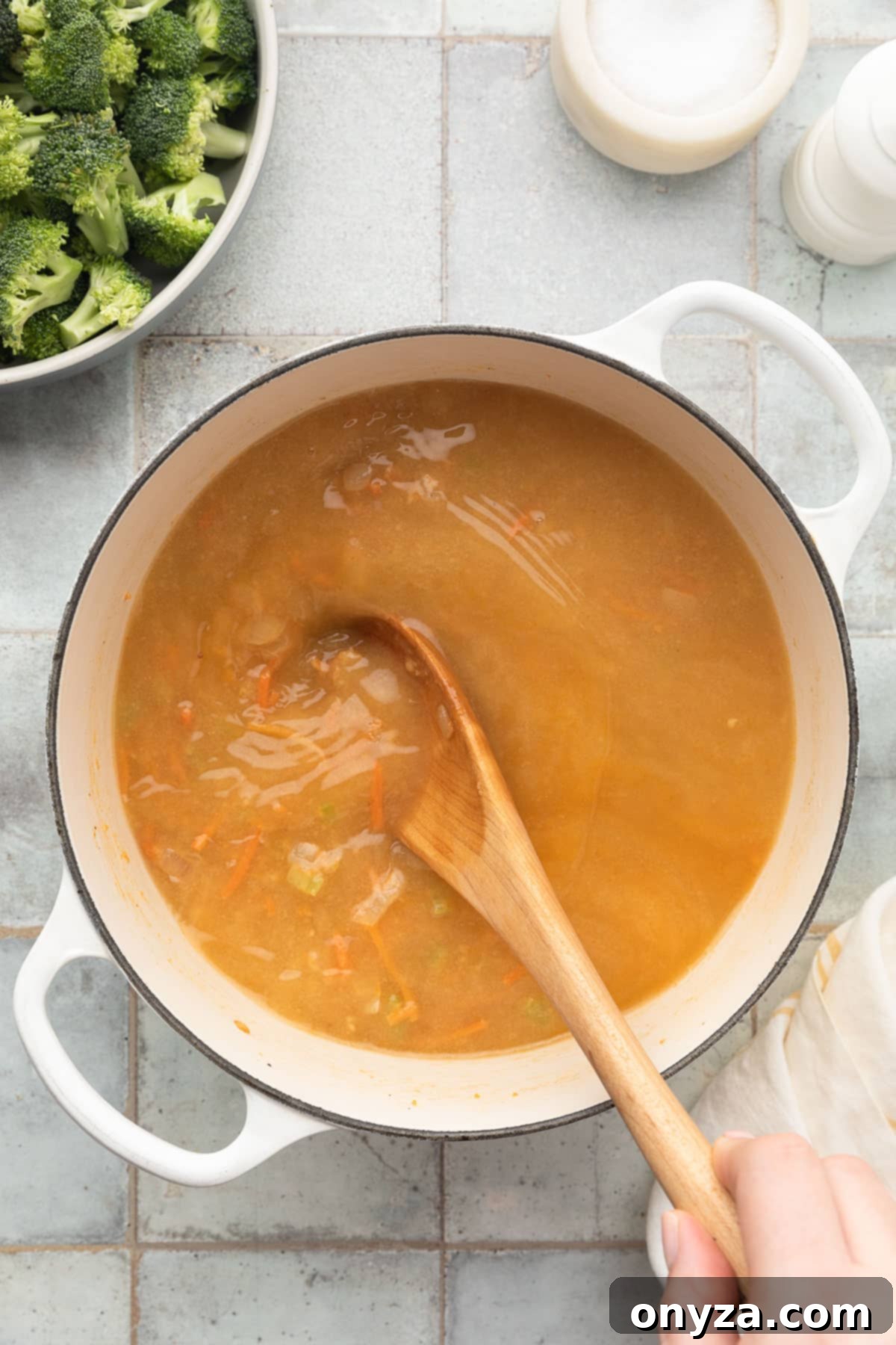 stirring chicken stock into sauteed vegetables in a white enameled cast iron dutch oven with a wooden spoon