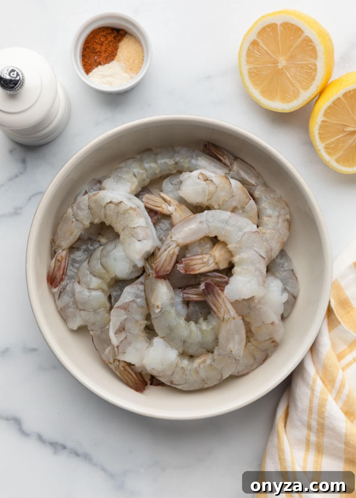 Crispy Air Fryer Shrimp 4 raw, peeled, deveined shrimp in an ivory ceramic bowl on a white marble surface next to lemon wedges, a pepper mill, a bowl of ground spices, and a white and yellow striped napkin