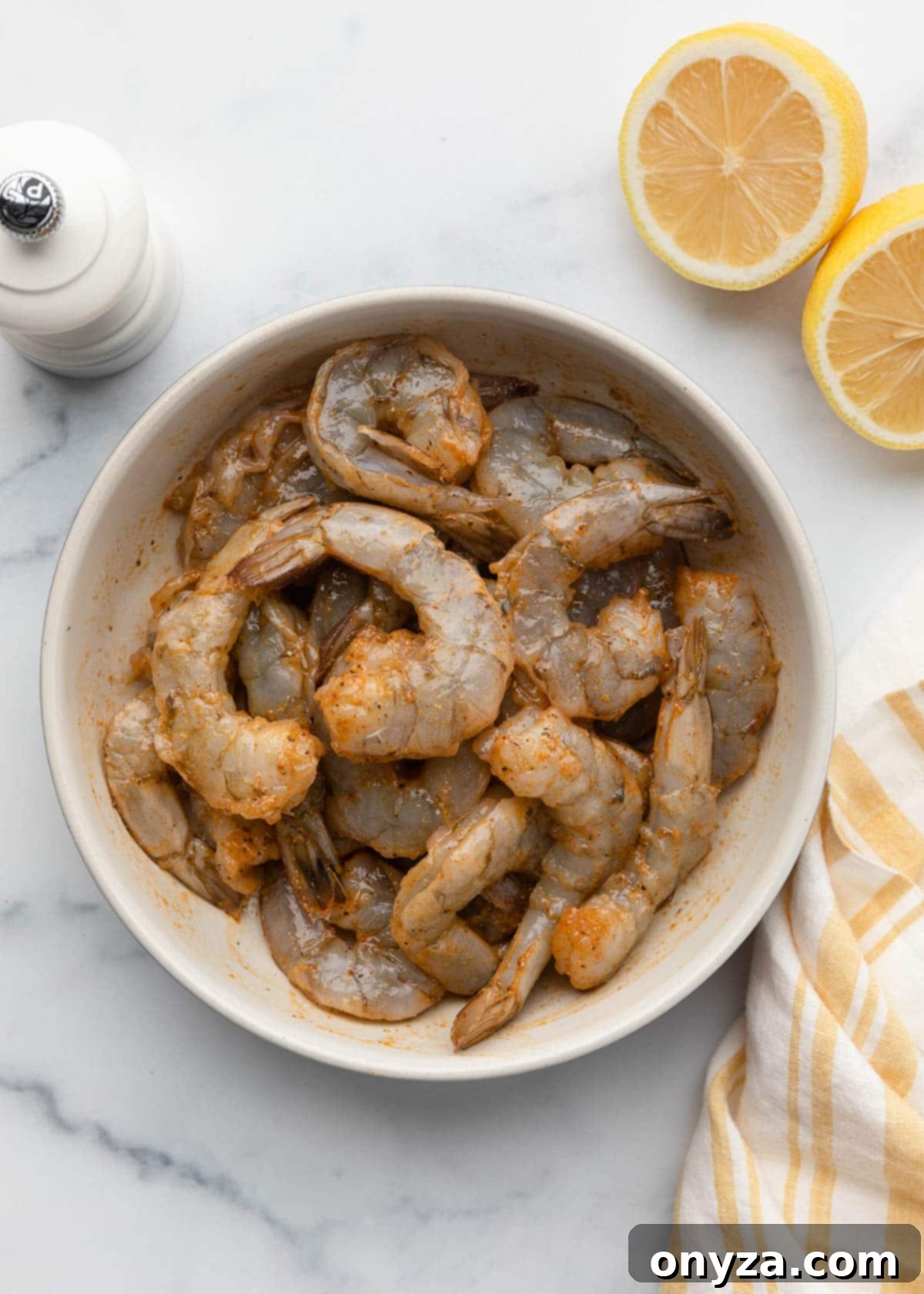 Crispy Air Fryer Shrimp 7 raw shrimp tossed with olive oil and spices in a bowl on a white marble board next to a halved lemon