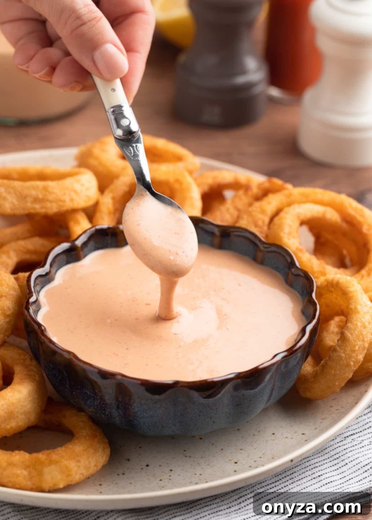 A spoonful of creamy Comeback Sauce drizzling into a blue scalloped bowl filled with the sauce, on a platter with golden onion rings, showcasing its rich texture.