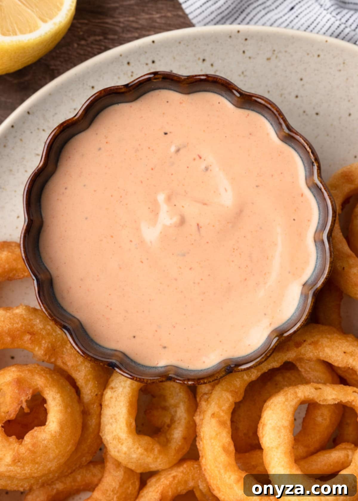 An overhead view of creamy Comeback Sauce in an elegant blue scalloped bowl, surrounded by a tempting platter of golden-brown onion rings, ready for dipping.