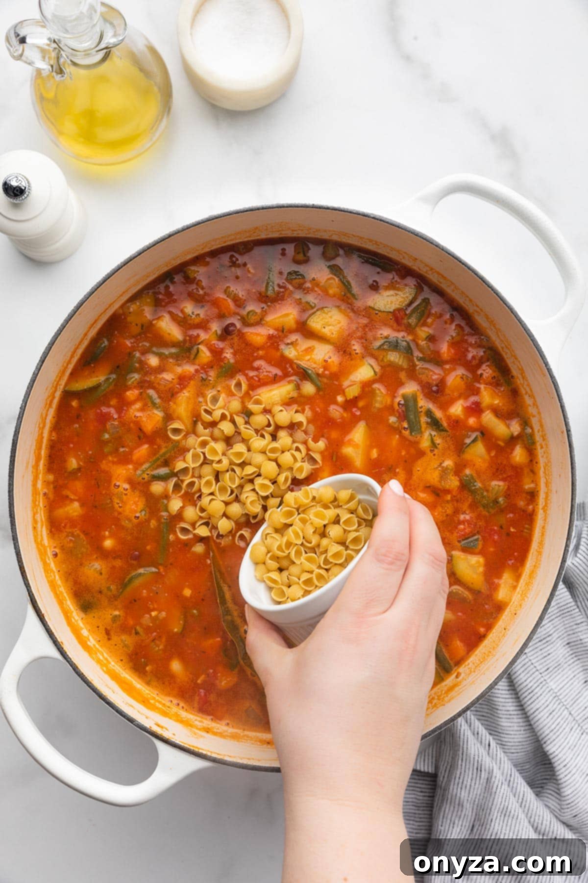 Pouring dry mini shell pasta from a small white ramekin into a pot of simmering Minestrone Soup.