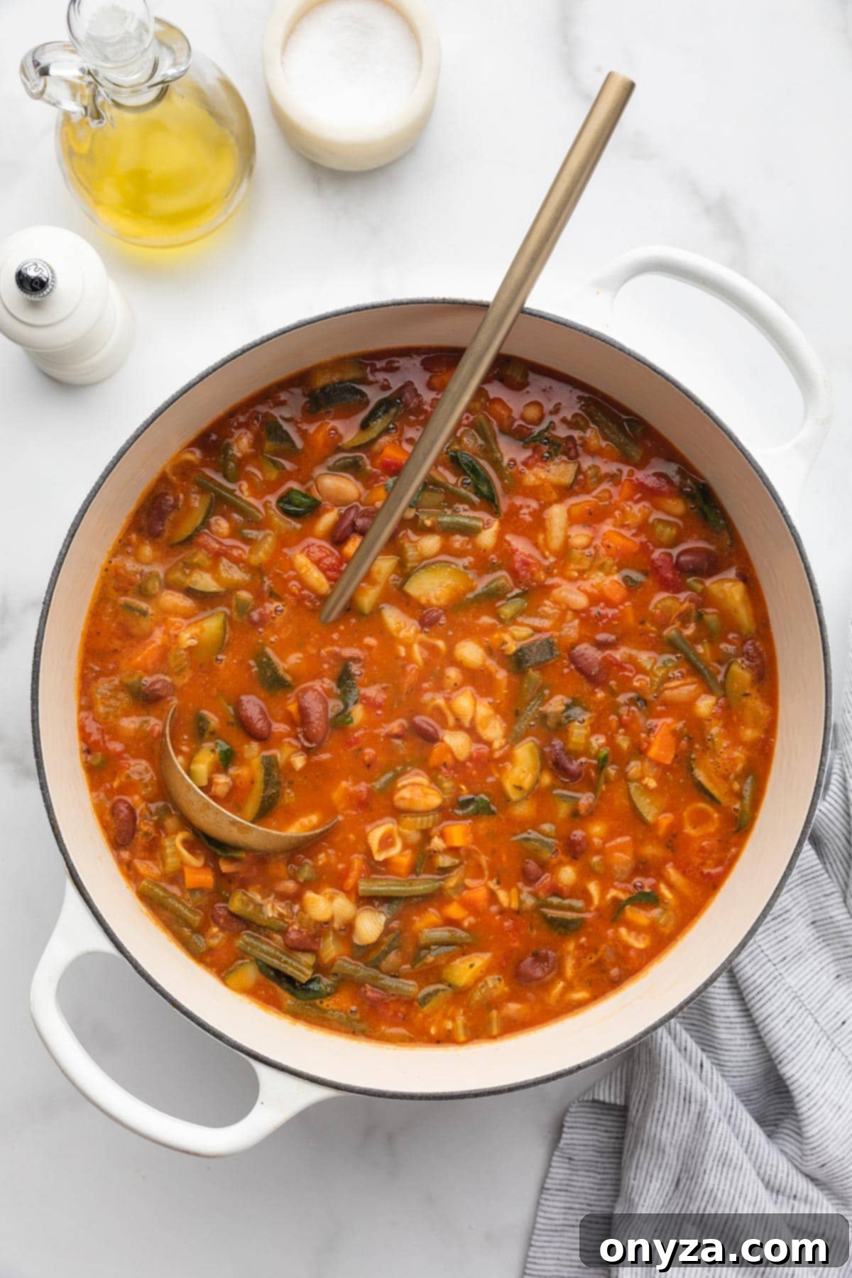 An overhead view of a rich, cooked Minestrone Soup in a white enameled cast iron Dutch oven, with a gold ladle resting inside.