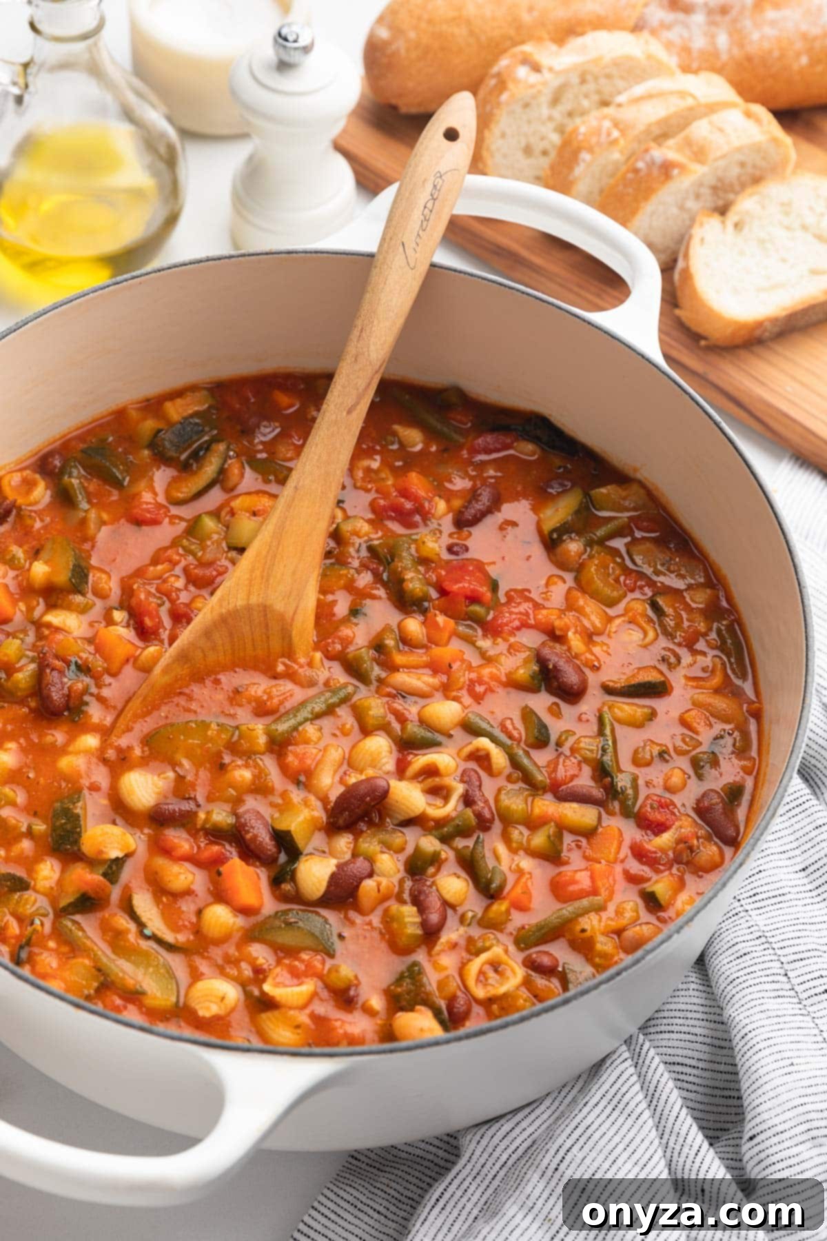A white enameled cast iron Dutch oven filled with Minestrone Soup and a wooden spoon, placed next to a cruet of olive oil and a wooden board with sliced Italian bread.