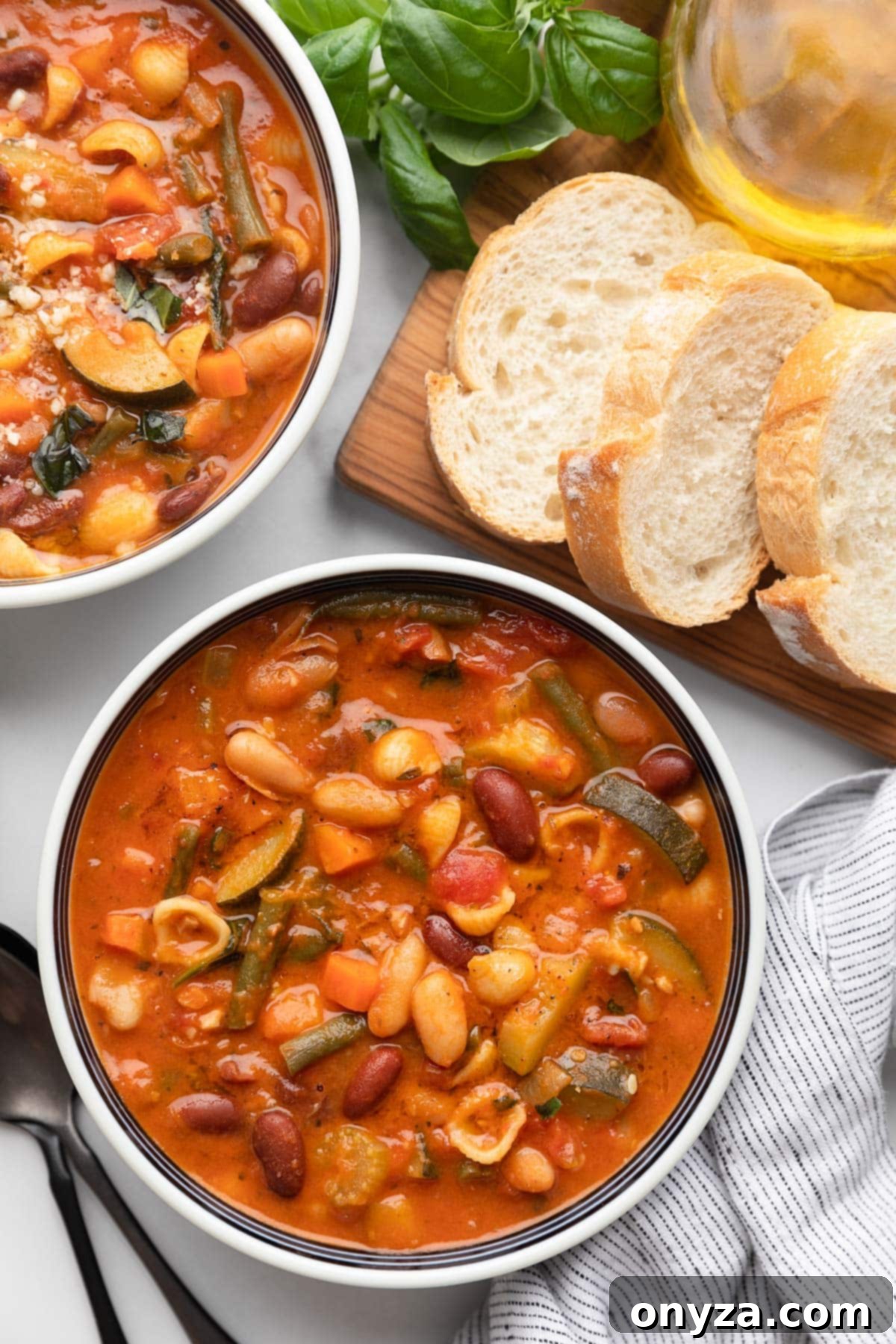 An overhead view of two inviting bowls of Minestrone Soup on a white marble board, accompanied by a bread board with freshly sliced Italian bread.