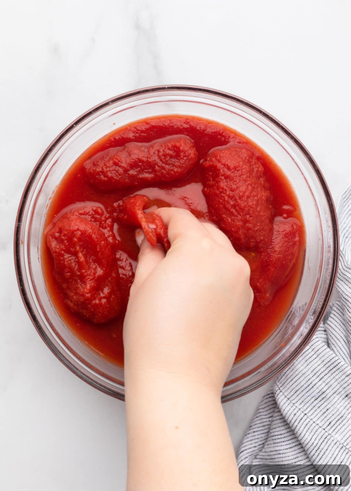 A hand crushing canned plum tomatoes in a clear glass bowl on a white marble surface.