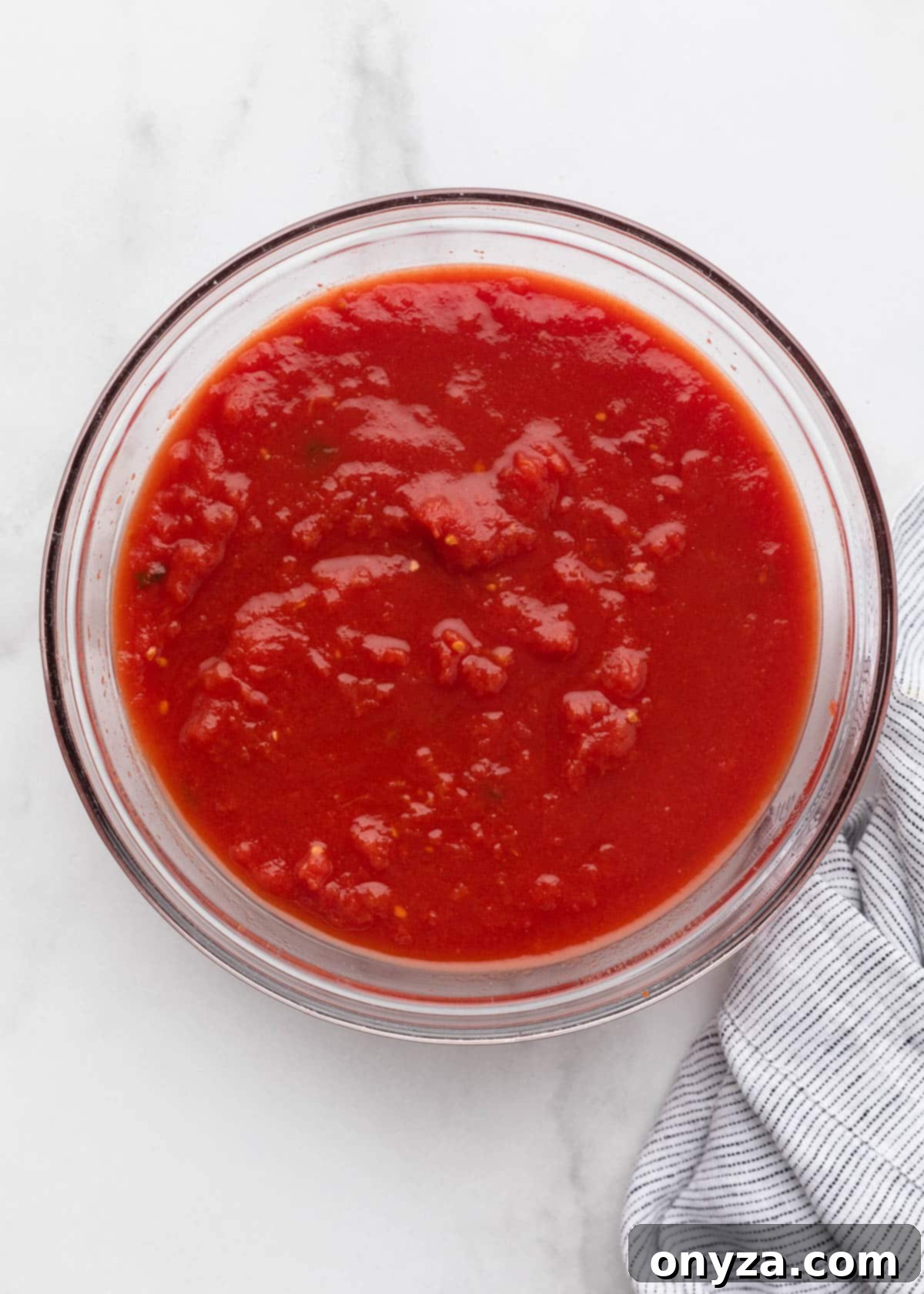 Finely hand-crushed plum tomatoes in a glass bowl, ready for cooking, on a white marble surface.