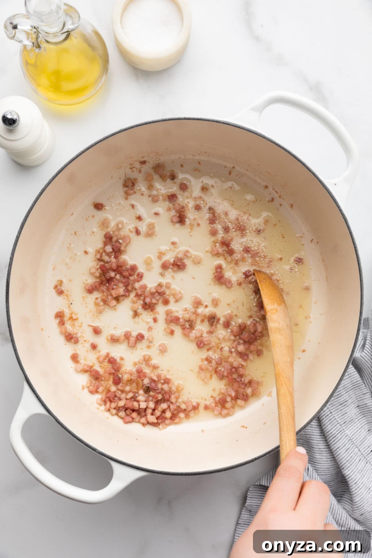 Diced pancetta gently frying in olive oil within a white enameled cast iron Dutch oven, being stirred with a wooden spoon.
