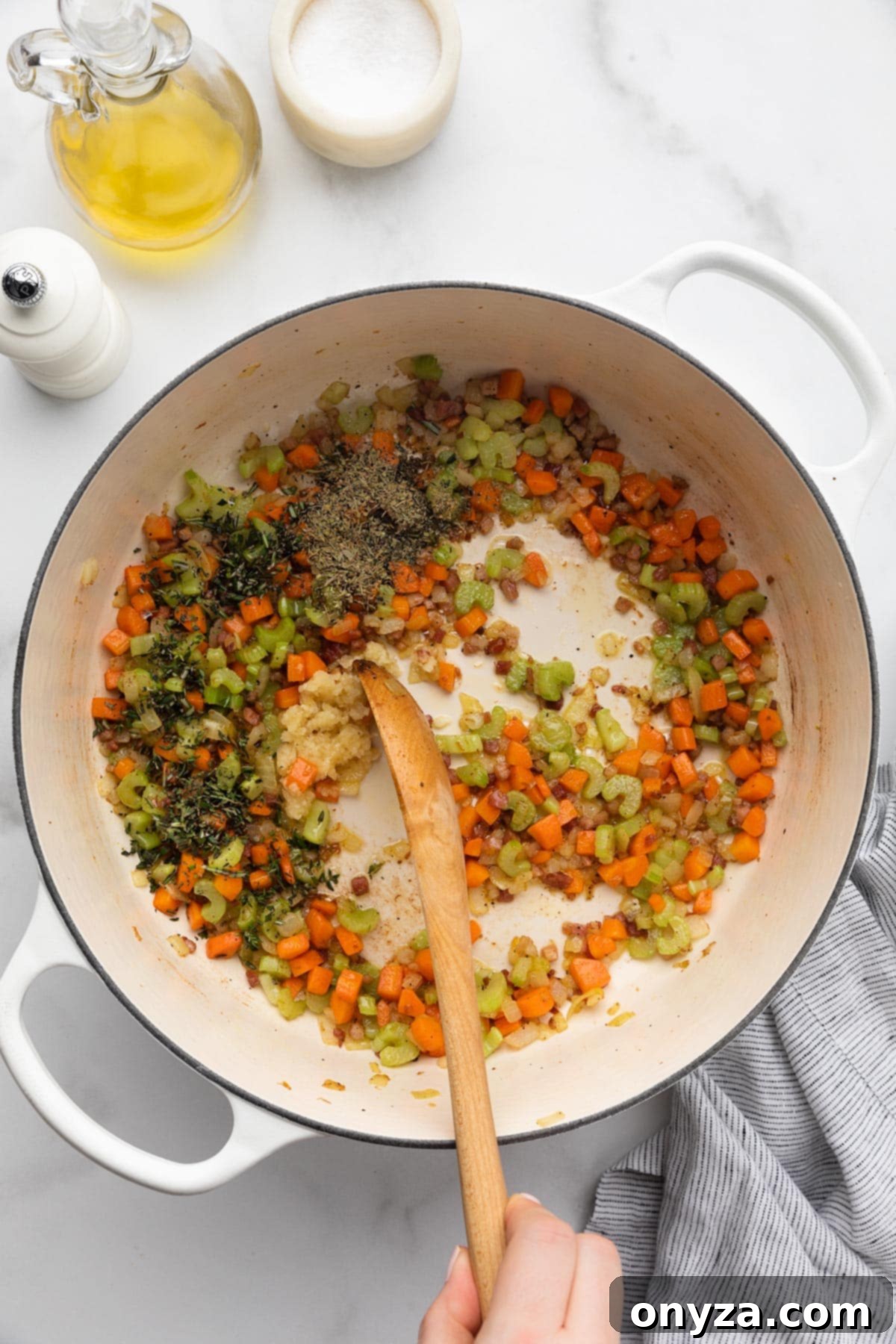 Minced garlic and finely chopped herbs being stirred into the cooked mirepoix in a white enameled cast iron Dutch oven with a wooden spoon.