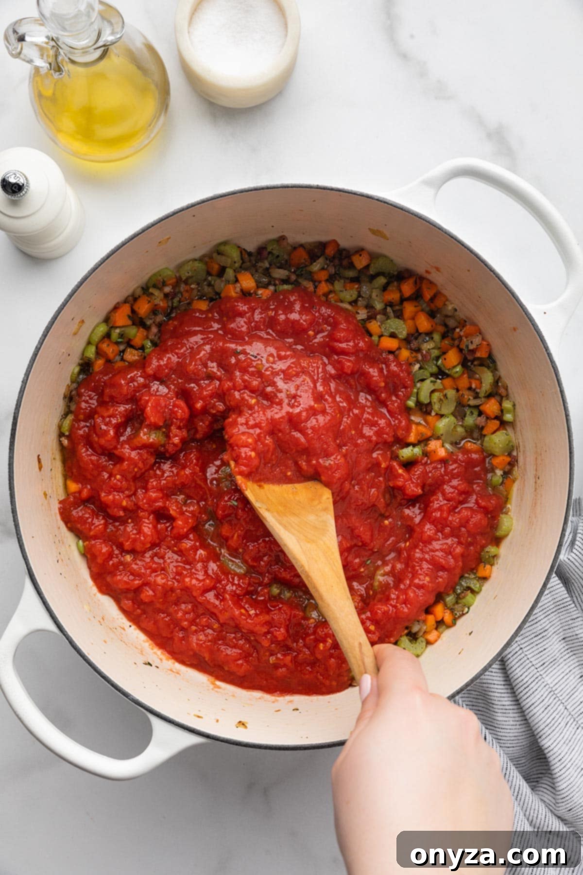 Crushed tomatoes being stirred into the cooked mirepoix in a white enameled cast iron Dutch oven using a wooden spoon.